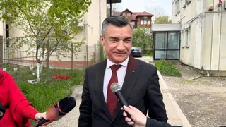 Un bărbat într-un costum negru și cravată roșie este intervievat de două microfoane în fața unor clădiri și copaci.
