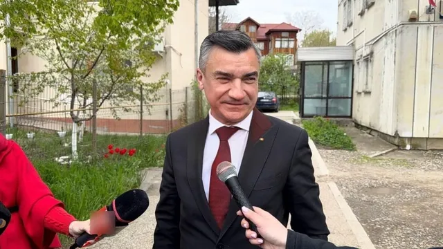 Un bărbat într-un costum negru și cravată roșie este intervievat de două microfoane în fața unor clădiri și copaci.