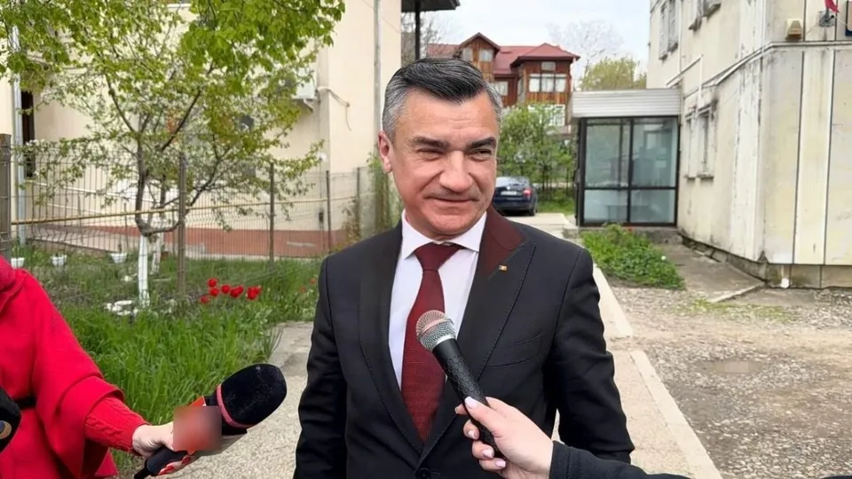 Un bărbat într-un costum negru și cravată roșie este intervievat de două microfoane în fața unor clădiri și copaci.