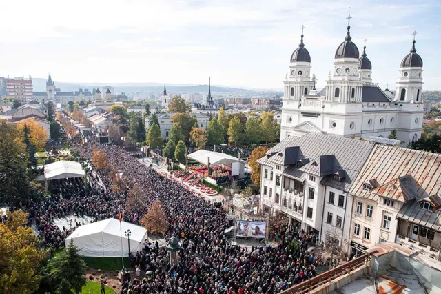 O mulțime mare de oameni este adunată într-o piață publică lângă o catedrală albă cu turne și câteva corturi albe.