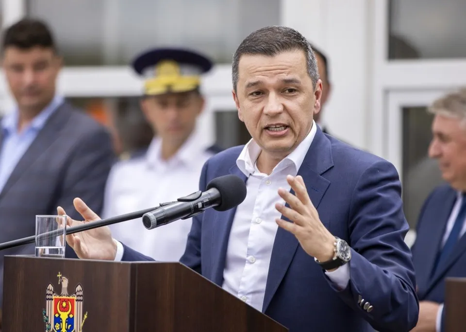 Un bărbat într-un costum albastru și cămașă albă vorbește la un microfon în fața unui podium maro.