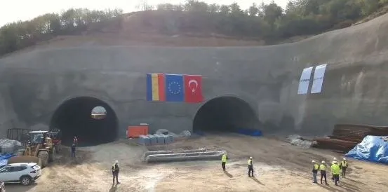Două tuneluri de beton sunt situate într-un deal, având trei steaguri suspendate între ele și mai mulți lucrători purtând veste reflectorizante.