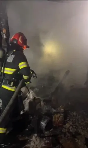 Un pompier cu echipament de protecție și cască roșie stă lângă un furtun într-un mediu plin de fum și resturi arse.