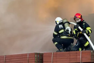 Doi pompieri cu echipament de protecție și cască stau ghemuiți pe un stivu de scânduri, ținând un furtun de apă sub cerul plin de fum.