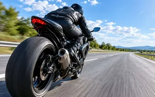O persoană purtând echipament de protecție negru conduce o motocicletă pe un drum asfaltat sub un cer cu nori albe.