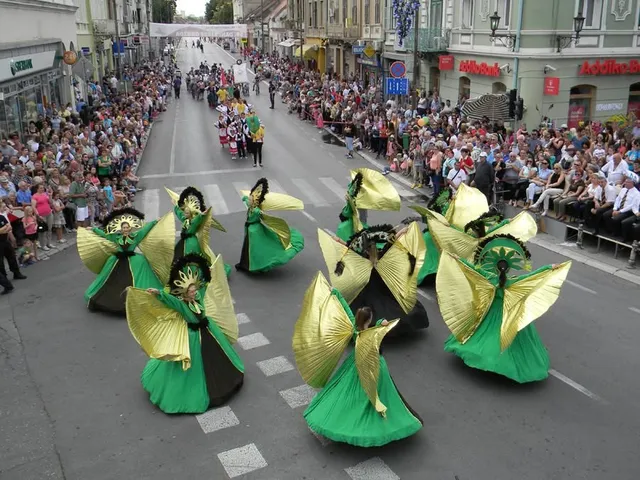 Oameni îmbrăcați în costume verzi și galbene cu aripi mari participă la un spectacol pe un drum asfaltat înconjurat de mulțimi.