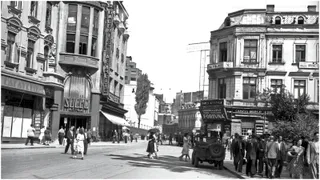 O stradă urbană aglomerată din trecut cu clădiri istorice, magazine cu vitrine, un automobil vechi pe partea dreaptă și mulți oameni în costume de epocă pe trotuar.