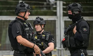 Trei bărbați în uniforme negre de polițist cu echipament tactical și căști de protecție stau în picioare lângă un gard metalic.