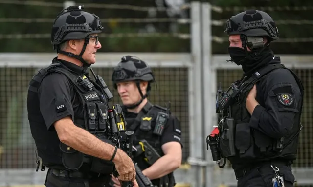 Trei bărbați în uniforme negre de polițist cu echipament tactical și căști de protecție stau în picioare lângă un gard metalic.