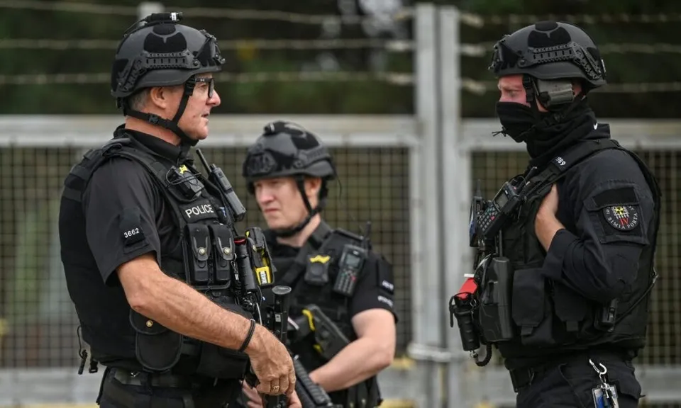 Trei bărbați în uniforme negre de polițist cu echipament tactical și căști de protecție stau în picioare lângă un gard metalic.