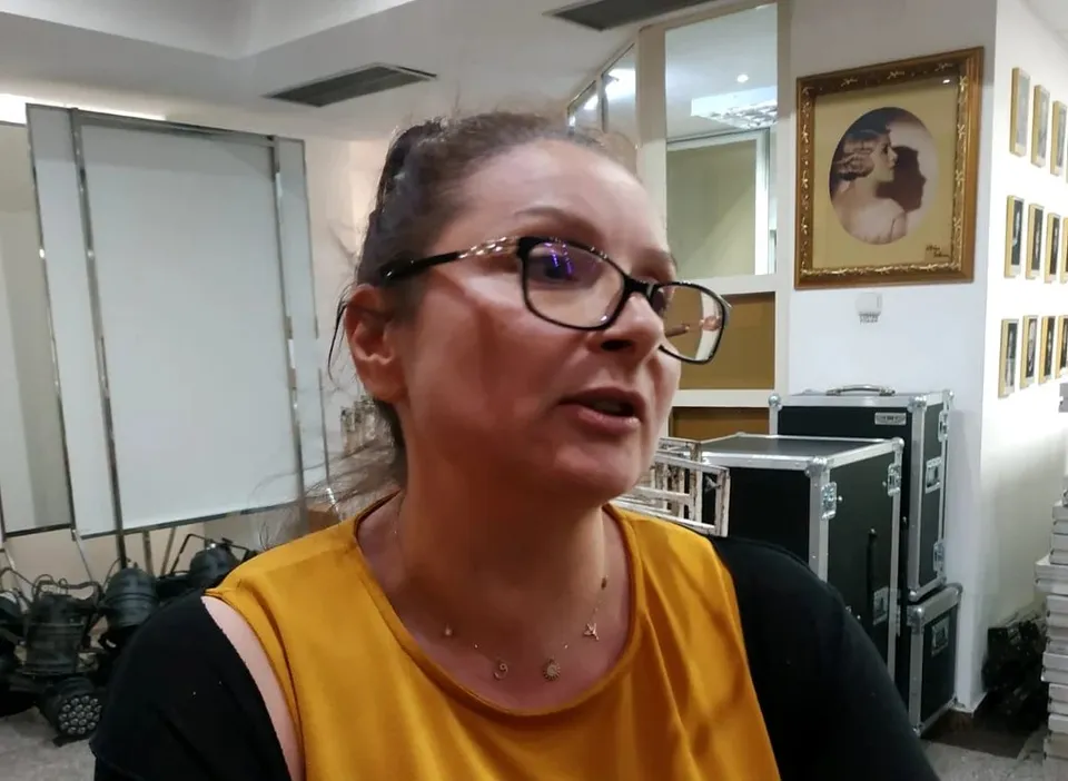 O femeie cu ochelari și bluză galbenă este surprinsă într-un cadru de interior lângă câteva cutii de transport negre și un tablou înrămat.