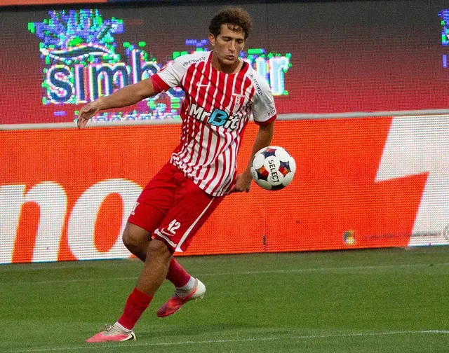 Un fotbalist într-un echipament alb cu dungi roșii și pantaloni roșii controlează un balon alb cu motive colorate pe un teren de fotbal.