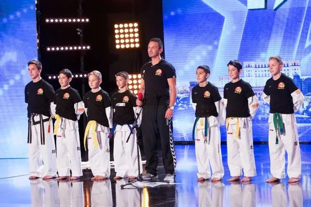 Un grup de băieți în echipamente de taekwon-do stau în linie pe o scenă lângă un bărbat care ține un microfon.