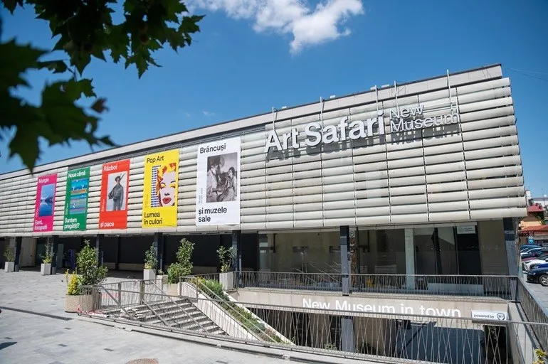O clădire modernă cu fațadă gri metalică prezintă diverse afișe colorate verticale și inscripția Art Safari New Museum.