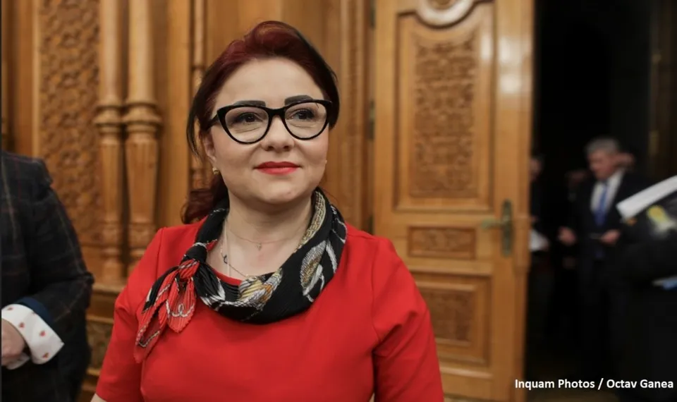 O femeie cu ochelari negri, purtând o bluză roșie și o eșarfă modelată, privește spre lateral într-un decor cu uși de lemn sculptate.