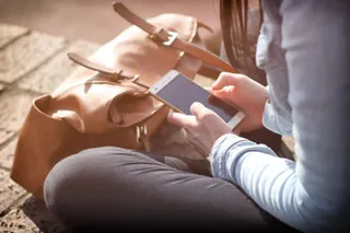 O persoană cu părul lung, purtând o cămașă albastră și pantaloni închiși, ține un telefon mobil în mâini lângă un rucsac maro.