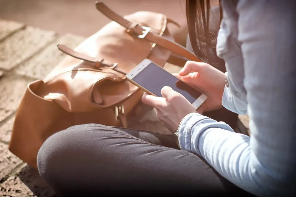 O persoană cu părul lung, purtând o cămașă albastră și pantaloni închiși, ține un telefon mobil în mâini lângă un rucsac maro.