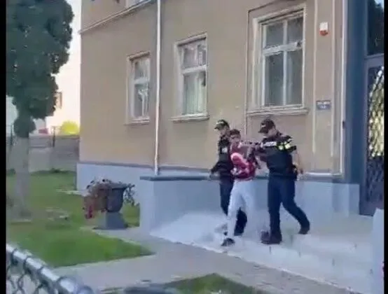 Trei persoane, dintre care două poartă uniforme închise la culoare, se află pe o treaptă de beton în fața unei clădiri cu ferestre albe.