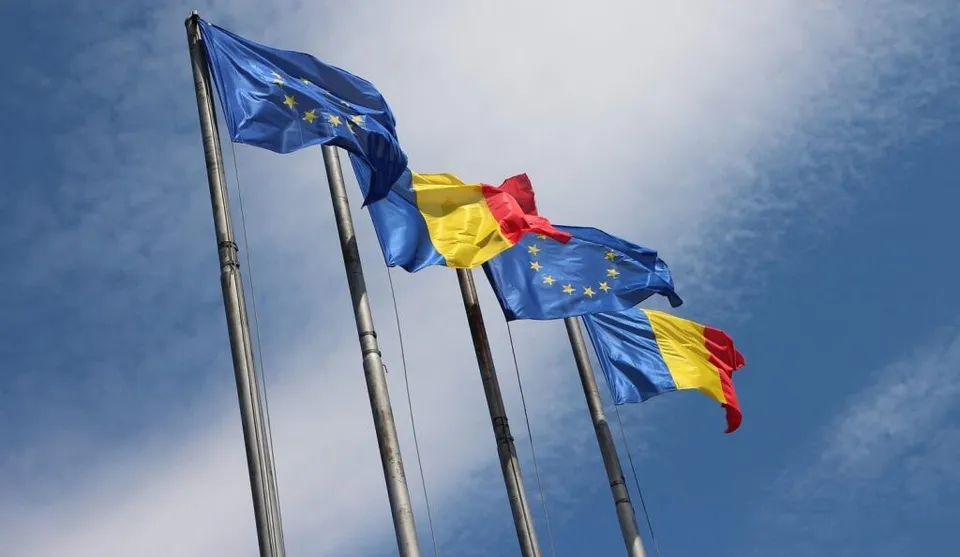 Patru steaguri, inclusiv steagurile Uniunii Europene și ale României, flutură pe catarge sub un cer albastru cu nori subțiri.
