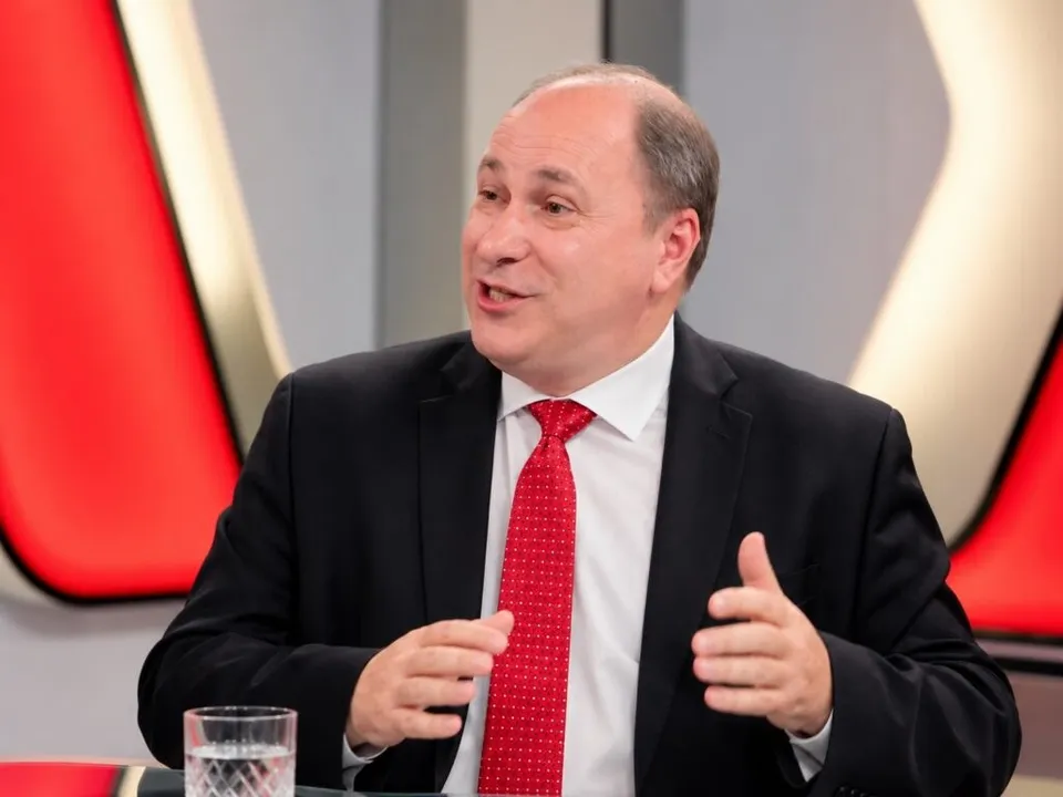 Un bărbat cu sacou negru, cămașă albă și cravată roată stă la o masă lângă un pahar cu apă transparentă.