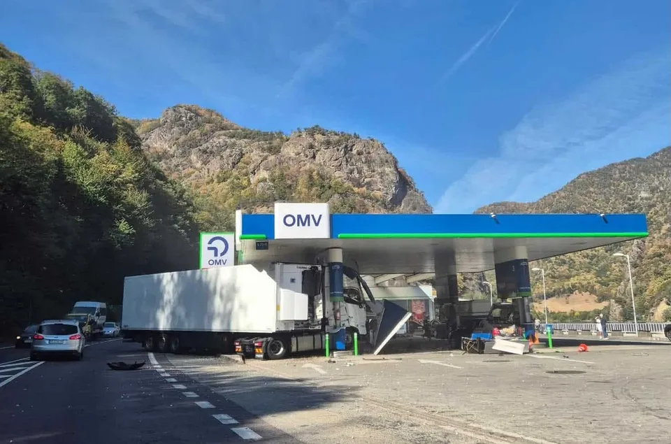 Un autocamion alb este poziționat lângă o stație de alimentare OMV, sub o canopelă albastră și verde, într-o zonă montană cu vegetație densă.