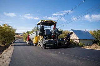 O mașină de asfaltat circulă pe un drum nou asfaltat lângă o casă și sub linii electrice sub un cer senin.