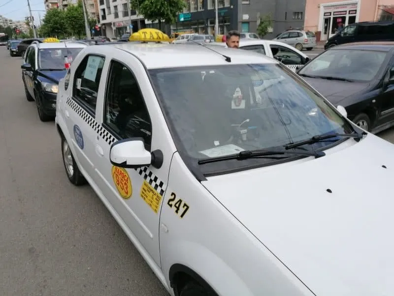 Un taxi alb cu numărul 247 și un semn galben pe plafon este parcat pe o stradă lângă alte mașini.
