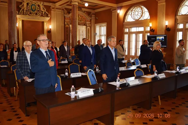 Un grup de bărbați în costume și o femeie stau în picioare în fața unor mese de conferință într-o sală cu coloane și ferestre înalte.