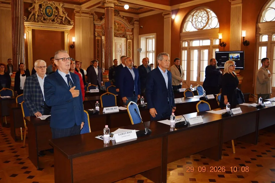 Un grup de bărbați în costume și o femeie stau în picioare în fața unor mese de conferință într-o sală cu coloane și ferestre înalte.