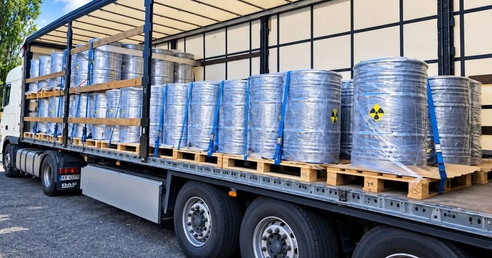 Containere cilindrice metalice învelite în folie transparentă sunt așezate pe paleți de lemn în interiorul unui camion cu platformă deschisă.