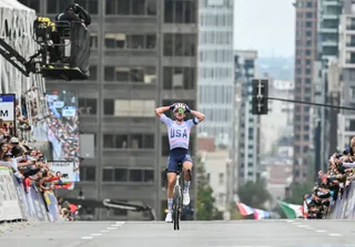 Un biciclist cu echipament albastru și alb cu inscripția USA ridică brațele în timp ce pedalează pe o stradă urbană.