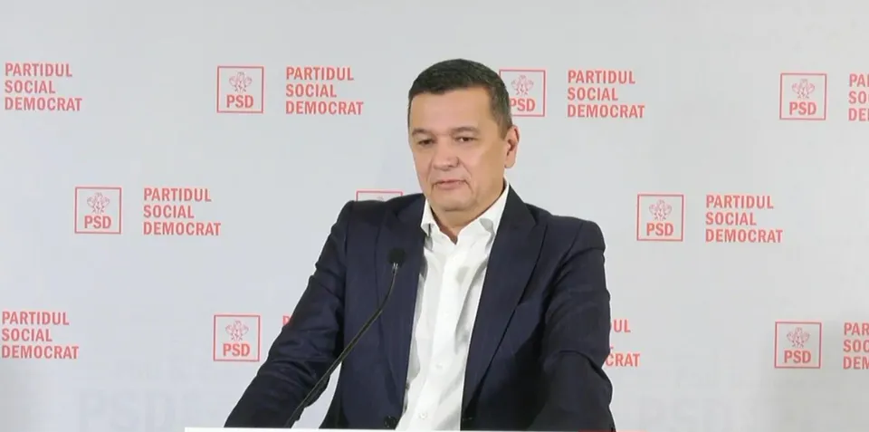 Un bărbat cu sacou închis și cămașă albă stă în fața unui fundal alb cu logouri roșii repetitive.
