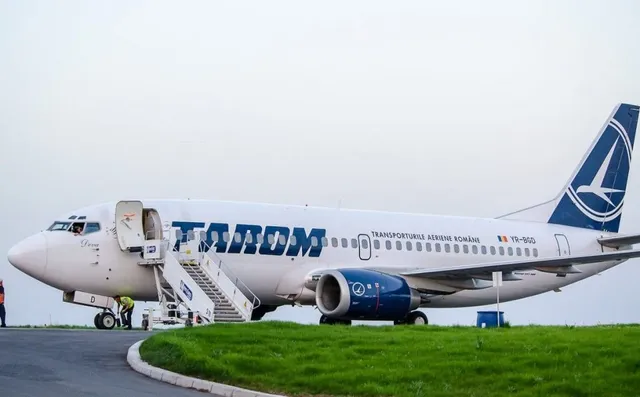 Un avion alb cu inscripția TAROM și un motor albastru sunt parcate pe o pista lângă o zonă cu iarba verde.