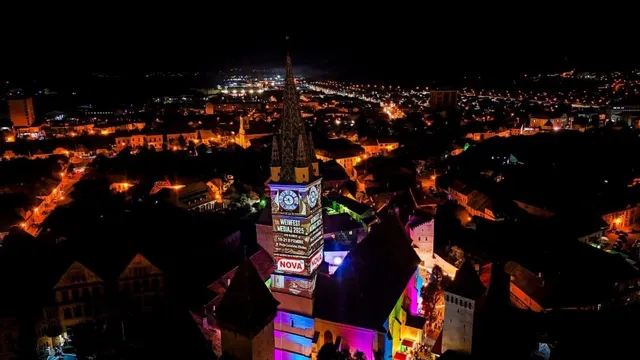 O vedere de noapte asupra unui oraș cu clădiri iluminate și un turn înalt cu un ceas colorat în centru.