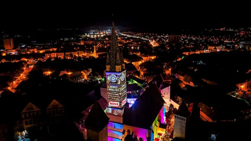 O vedere de noapte asupra unui oraș cu clădiri iluminate și un turn înalt cu un ceas colorat în centru.