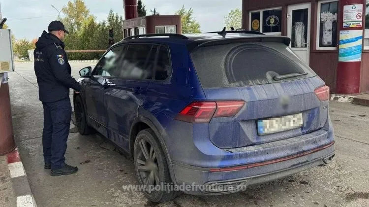 Un agent de poliție în uniformă întunecată stă lângă o mașină de culoare albastru închis aflată pe o platformă de transport.