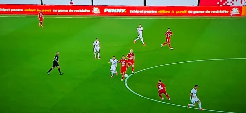Jucători de fotbal în echipamente albastre și roșii sunt dispersați pe un teren de fotbal verde sub lumină artificială.