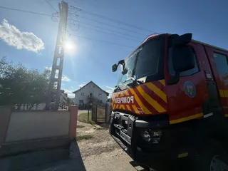 Un camion de pompieri roșu cu dungi galbene este parcat pe un drum de țară lângă un stâlp electric și o casă.