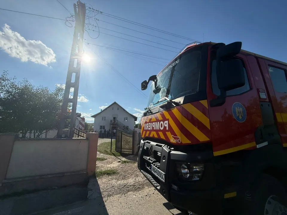 Un camion de pompieri roșu cu dungi galbene este parcat pe un drum de țară lângă un stâlp electric și o casă.