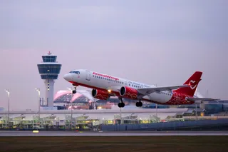 O aeronavă albă cu accente roșii și inscripția AnimaWings decolează de pe o pistă de lângă un turn de control și terminale de aeroport.