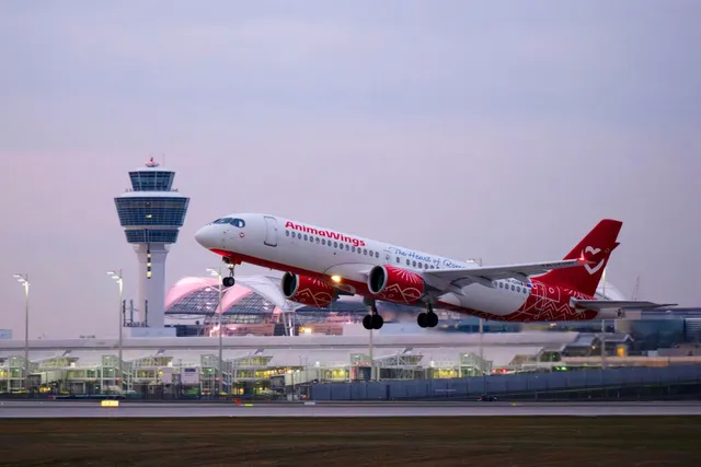 O aeronavă albă cu accente roșii și inscripția AnimaWings decolează de pe o pistă de lângă un turn de control și terminale de aeroport.