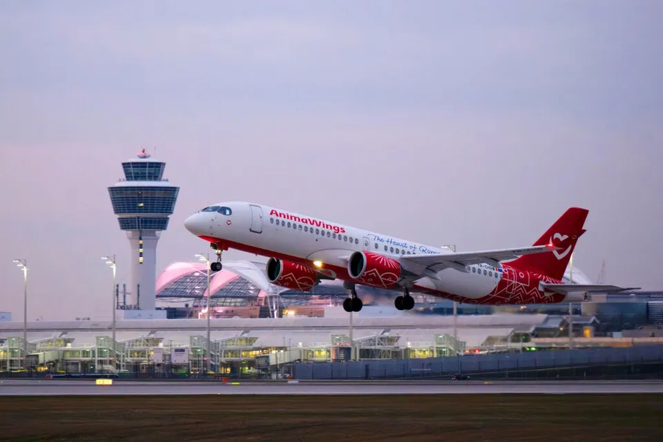 O aeronavă albă cu accente roșii și inscripția AnimaWings decolează de pe o pistă de lângă un turn de control și terminale de aeroport.