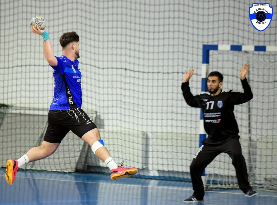 Un jucător de handbal în tricou albastru sare și lovește mingea, în timp ce portarul în negru ridică brațele la poartă.