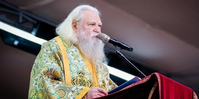 Un bărbat cu părul și barba albe, purtând o robă decorată cu motive aurii, vorbește la un microfon aflat deasupra unui podium roșu.