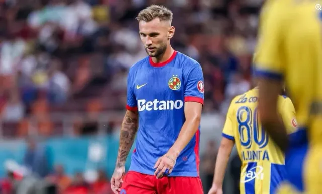 Un fotbalist cu tricou albastru și pantaloni roșii stă pe terenul de fotbal, privind în jos, în timp ce alți jucători sunt vizibili în fundal.