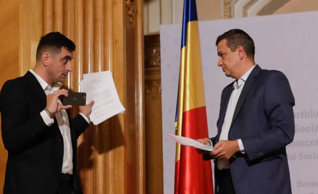 Un bărbat în costum negru ține un telefon și o foaie de hârtie, în timp ce un alt bărbat în costum albastru privește un document.