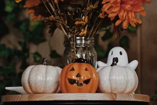 Un buchet de flori uscate și dovleci decorativi, inclusiv unul cu față de jack-o'-lantern și unul în formă de fantomă, stau pe o masă circulară.