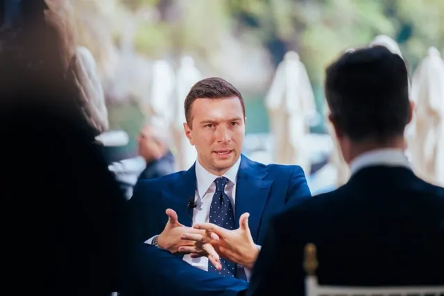 Un bărbat într-un costum albastru și cravată cu puncte vorbește cu alți doi bărbați în cadrul unui eveniment.