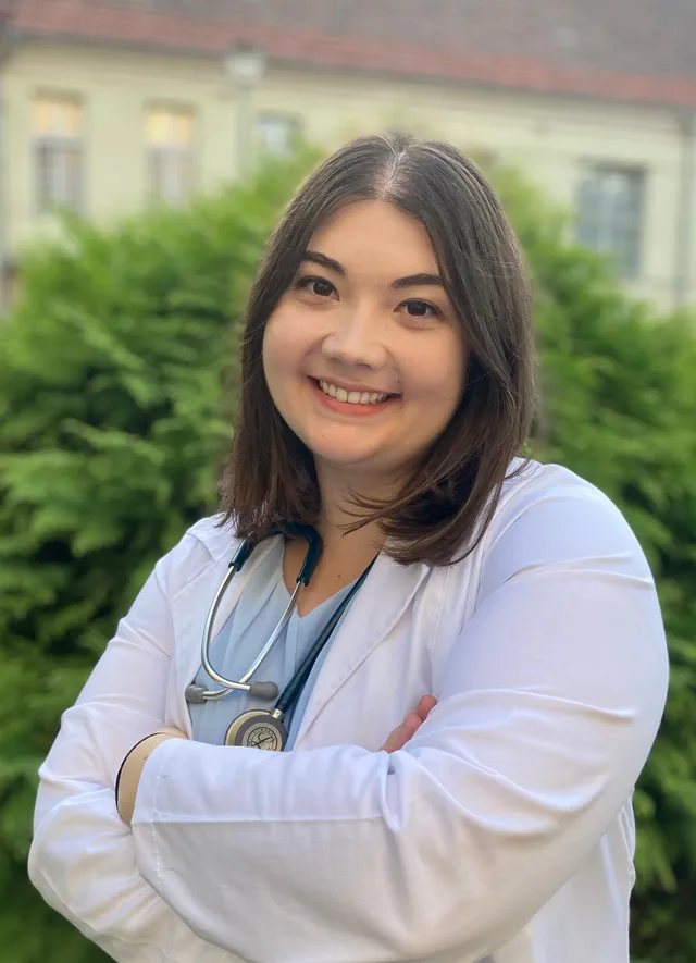 O femeie cu părul scurt și negru poartă o halat albă și un stetoscop în jurul gâtului, stând cu brațele încrucișate în fața unor plante verzi.