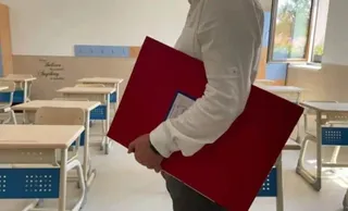 O persoană purtând o cămașă albă poartă un dosar roșu mare într-o sală de clasă cu bănci albe și scaune de culoare albastră.